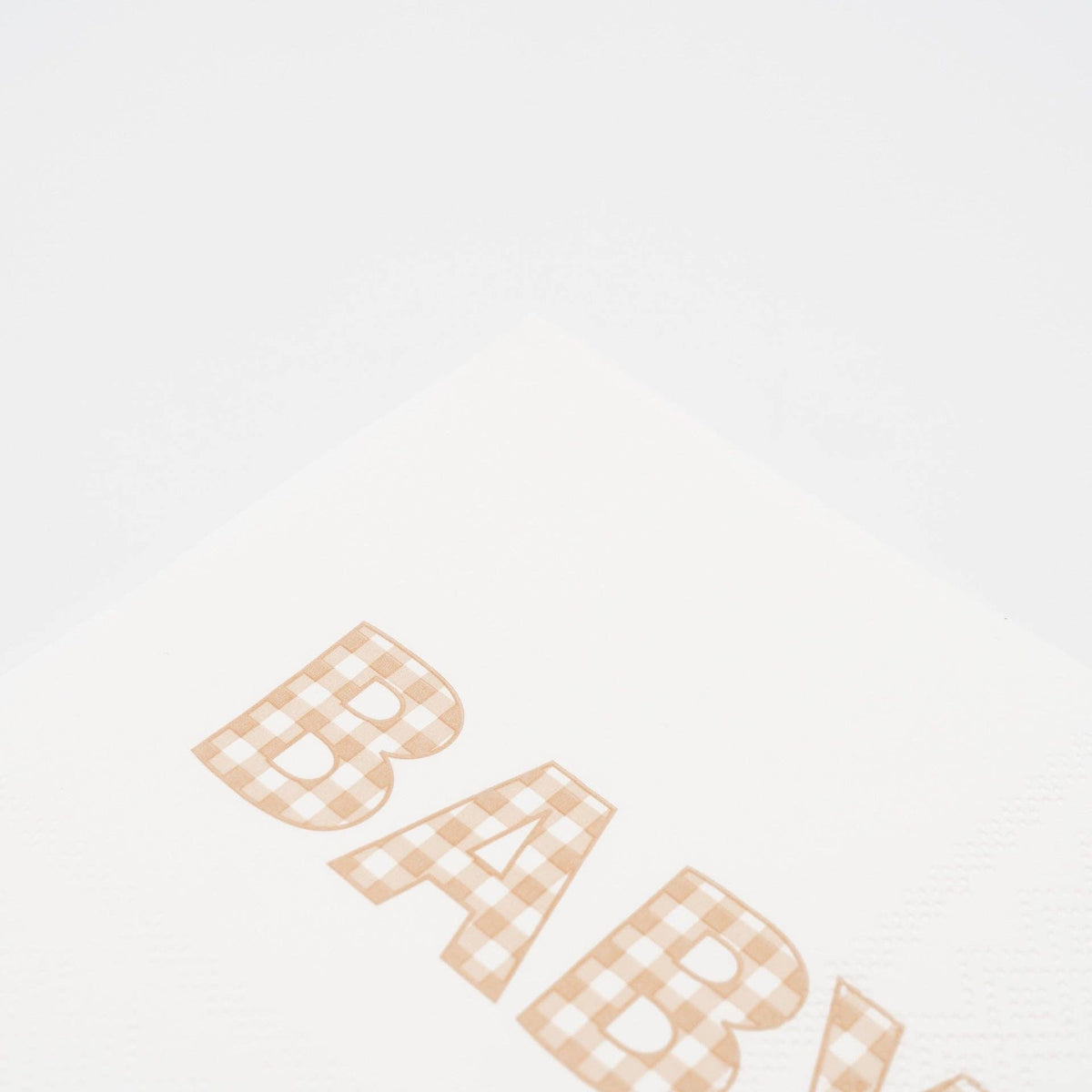 Close crop of the BABY lettering in tan gingham print on a white paper napkin