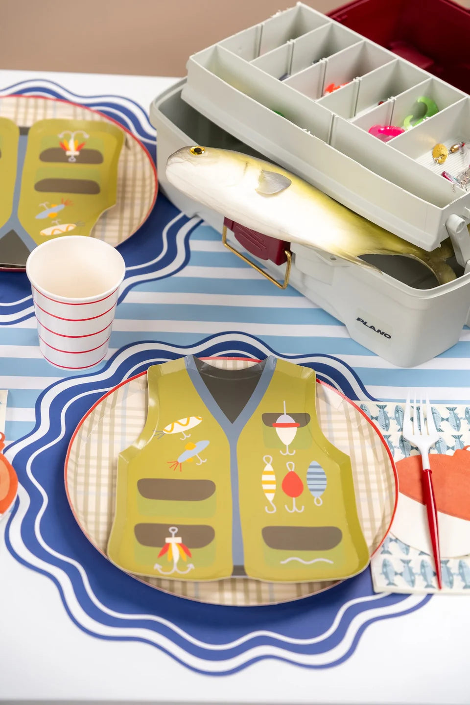 Tan plaid dinner plate as the base under a fishing vest shaped plate, with a striped cup, fish napkin, and tackle box.