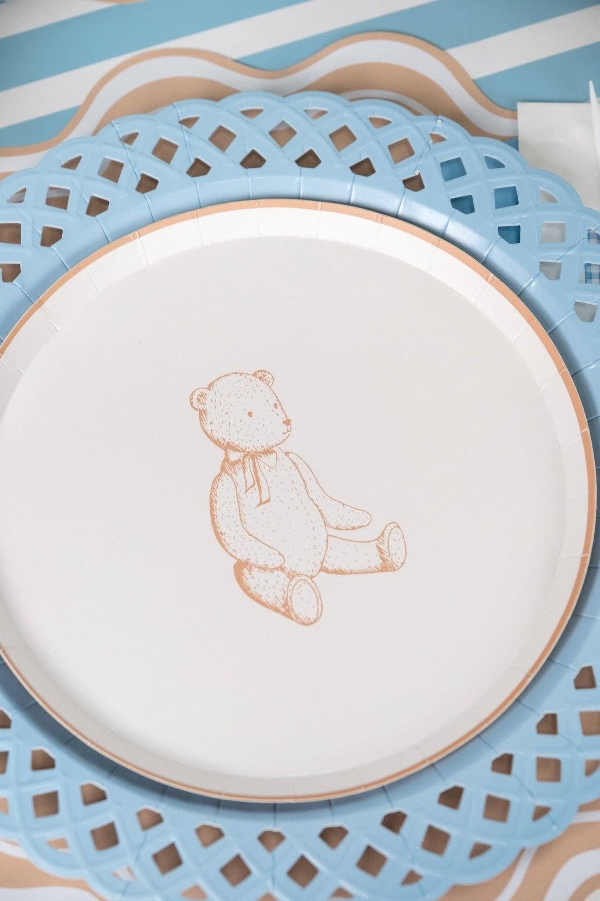 Close view of a tan teddy bear paper plate on a blue lattice charger over a blue striped runner