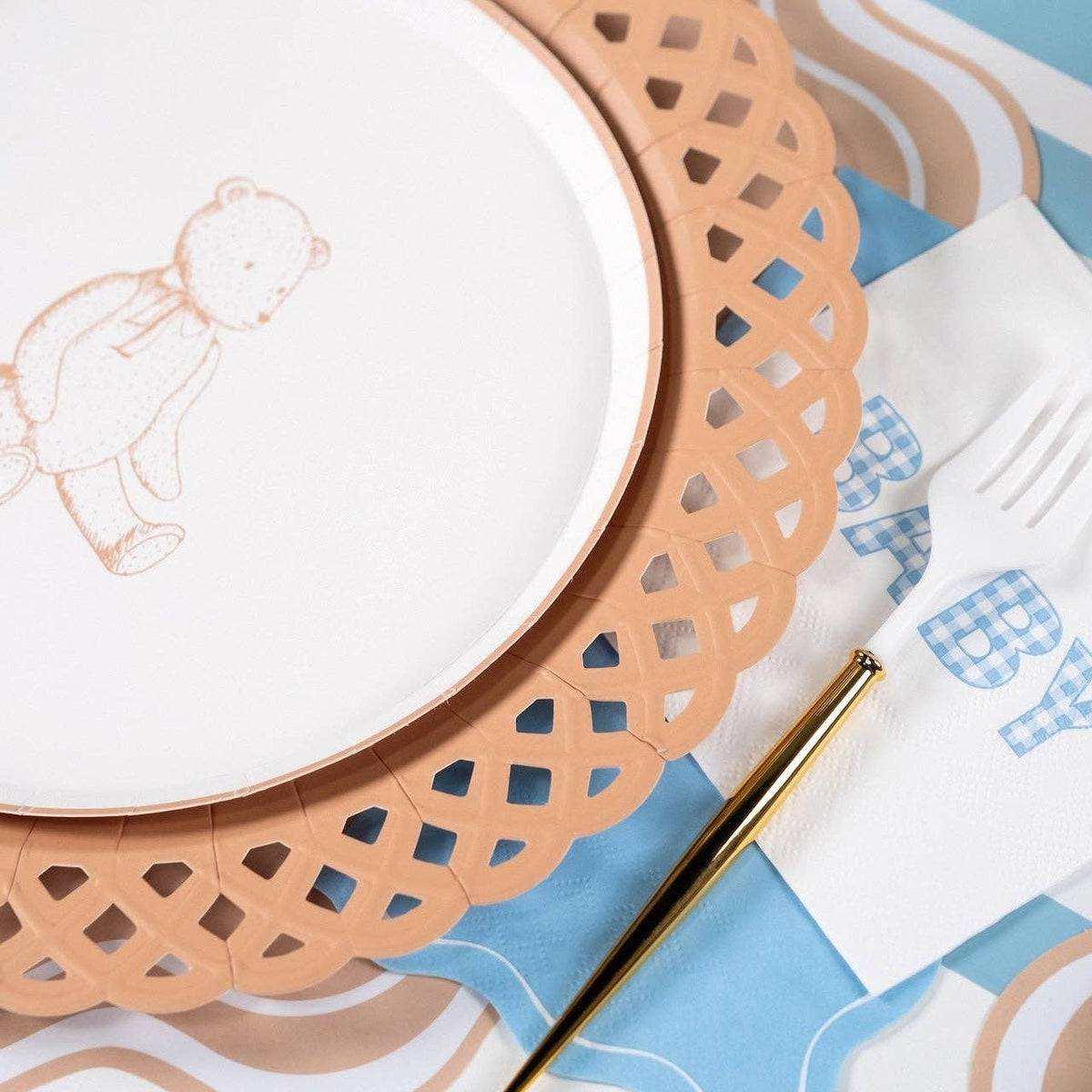 Edge of a tan teddy bear paper plate on a tan lattice charger beside a blue gingham BABY napkin