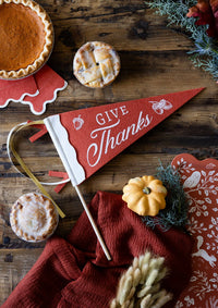 Styled Thanksgiving table overhead showing the Give Thanks pennant laid on a wood surface with pie, cookies, a rust napkin, and fall greenery.