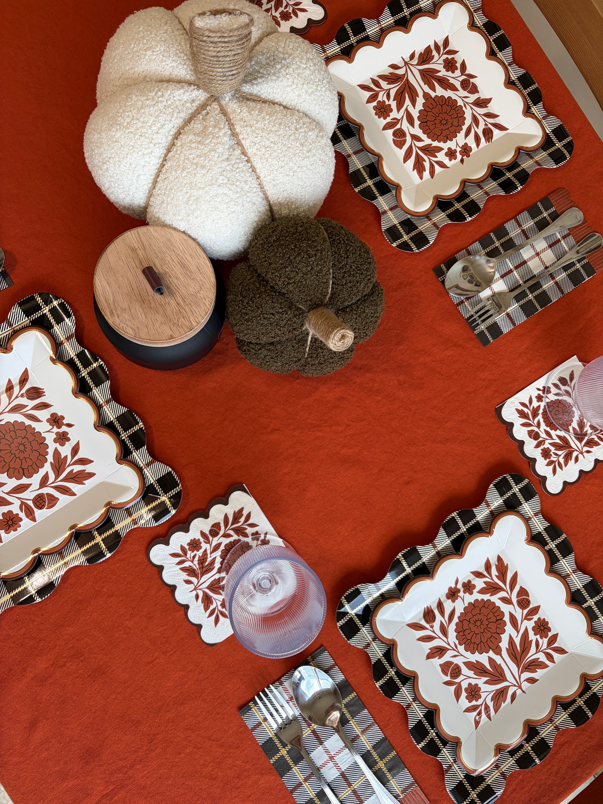 Overhead Thanksgiving tablescape with white and green knit pumpkins, a wood-lidded candle, rust floral square plates, folded plaid napkins with gold flatware, and a pink glass on an orange tablecloth