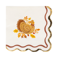 Cream Thanksgiving cocktail napkin with a detailed brown turkey illustration surrounded by fall leaves and acorns, gold foil wavy accent, and wavy orange edges, overhead flat-lay on gray background.