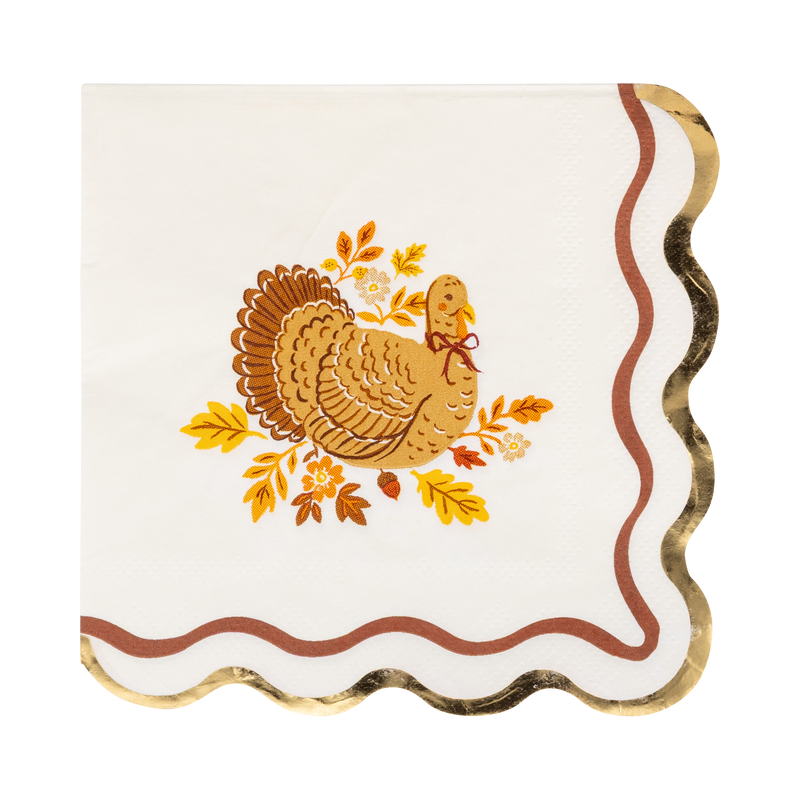 Cream Thanksgiving cocktail napkin with a detailed brown turkey illustration surrounded by fall leaves and acorns, gold foil wavy accent, and wavy orange edges, overhead flat-lay on gray background.
