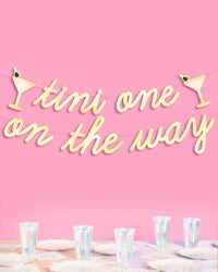 Tini One on the Way gold foil banner hung above baby shower table with cups and napkins