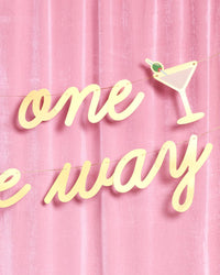 Close-up of gold foil script and martini glass icon on Tini One on the Way banner against pink velvet