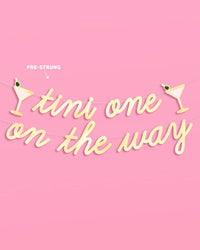 Tini One on the Way gold foil banner pre-strung on gold cords with martini glass icons on pink background
