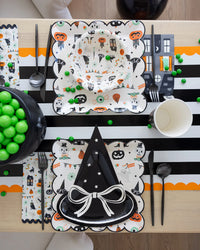 Halloween-themed table setting with decorative plates, cups, and utensils on a black and white striped tablecloth.