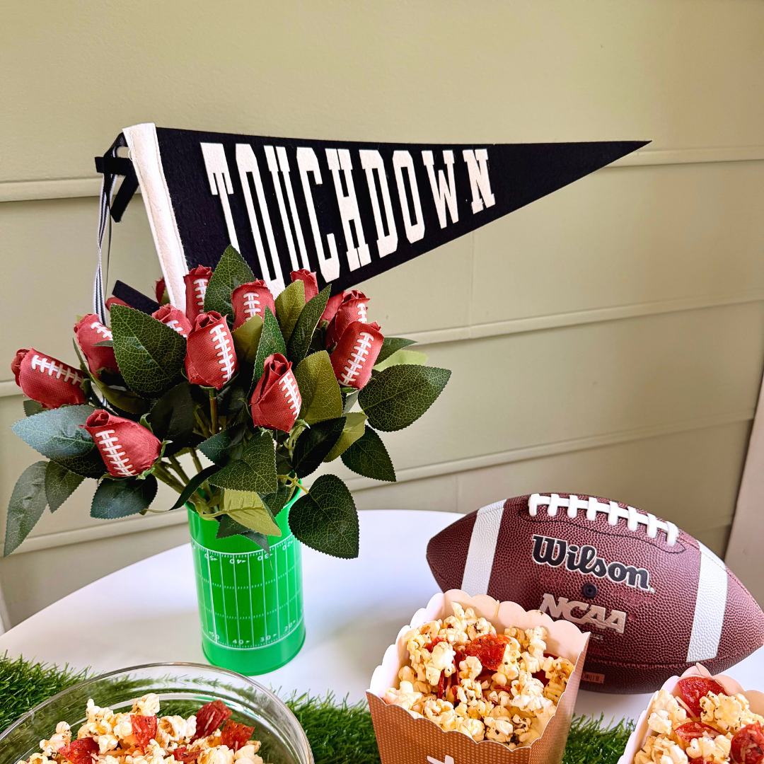 A football-themed party centerpiece on a white table. A bright green vase holds a bouquet of artificial red roses, each painted with white laces to look like mini footballs. A black and white felt pennant flag reading "TOUCHDOWN" sticks out of the arrangement. A Wilson NCAA leather football and a dish of popcorn sit next to the vase against a light green paneled wall.