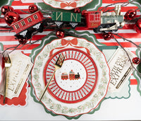 Place setting with a toy-train plate on the wreath dinner plate and green charger, with a "North Pole" banner and red ornaments.