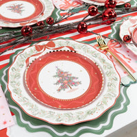 Place setting with the tree dessert plate on a red scalloped plate and evergreen-rim plate, with red ornaments and a striped runner.