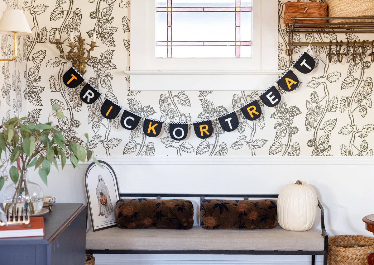 "Trick or Treat" felt banner hung across a wall above a bench in a styled entryway. Black felt flags with alternating orange and cream letters drape in a gentle curve beneath a stained-glass window. Botanical-print wallpaper on both sides. Below the banner: a gray upholstered bench with brown floral cushions, a white pumpkin, and an arched ghost art print leaning against the backrest. Green eucalyptus branch in a glass vase on a side cabinet at left.