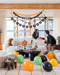 Wide shot of a living room decorated for a kids' Halloween party. The "Trick or Treat" felt banner hangs across a large window behind a white bouclé sofa, with a layered orange-and-black scalloped garland above it and a Halloween character garland (die-cut icons: spider, bat, pumpkin, haunted house, black cat, witch, skull, cauldron) below. Two children in black jackets sit on the sofa. A black coffee table holds pumpkin-shaped plates, cupcakes, haunted house favor boxes, and a "Stay Spooky" pennant.