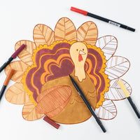 The coloring placemat with markers laid on top, showing the turkey partly colored in rust, burgundy, and orange.