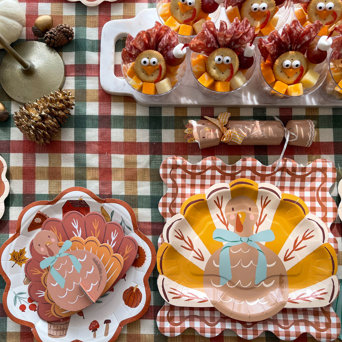 Turkey shaped paper plate with a blue bow on a scalloped gingham placemat, with a turkey party cracker and turkey snack cups behind it