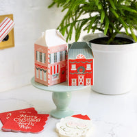 Two assembled Christmas cookie boxes displayed on a small cake stand, alongside matching paper goods.
