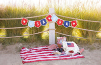 Wide shot of USA felt pennant banner displayed on beach rope fence at golden hour with American flag picnic blanket and basket below