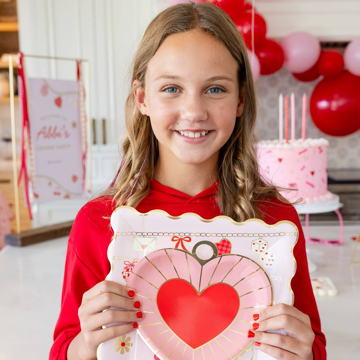 Smiling girl in red hoodie holding a pink scalloped Valentine charm plate at a Valentine-themed party.