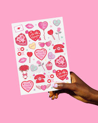 Hand holding Valentine's temporary tattoo sheet showing full range of vintage heart, cherub, and bow designs