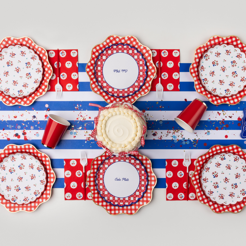 Overhead styled table with vintage floral dessert plates layered on red gingham dinner plates alongside cherry napkins, polka dot napkins, cake plates, red cups, confetti, and cake on blue striped table runner
