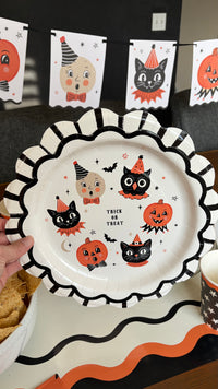 Vintage Halloween character paper plate with cats, owls, and pumpkins on a table layered with a black scalloped charger, star cups, and the character banner in the background