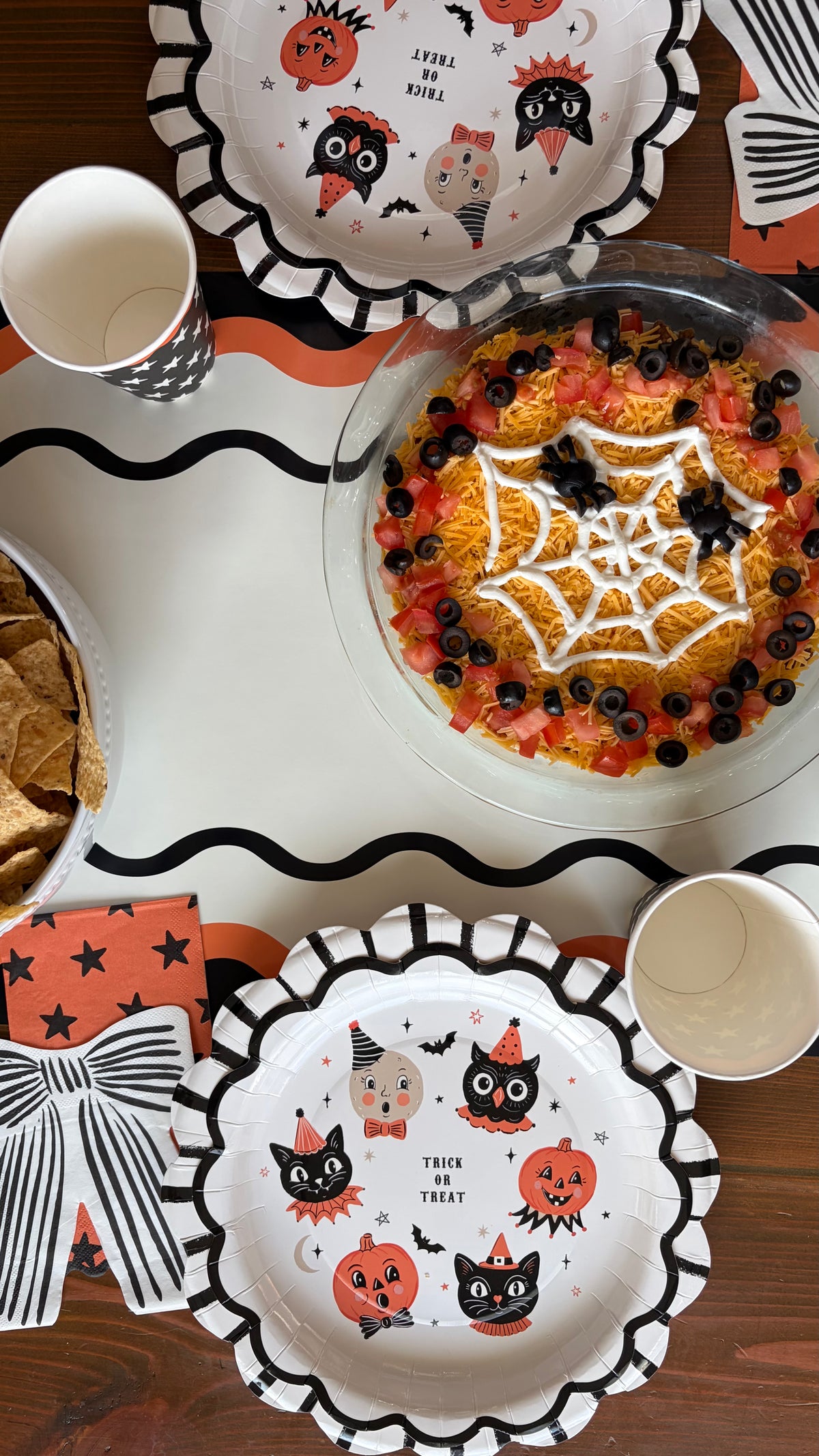 Overhead vintage Halloween table with character paper plates, a spiderweb taco dip, star cups, a black bow napkin, and orange and black ric-rac accents