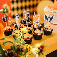 Tray of Halloween cupcakes with orange frosting and vintage character toppers on a yellow dessert table beside a cake
