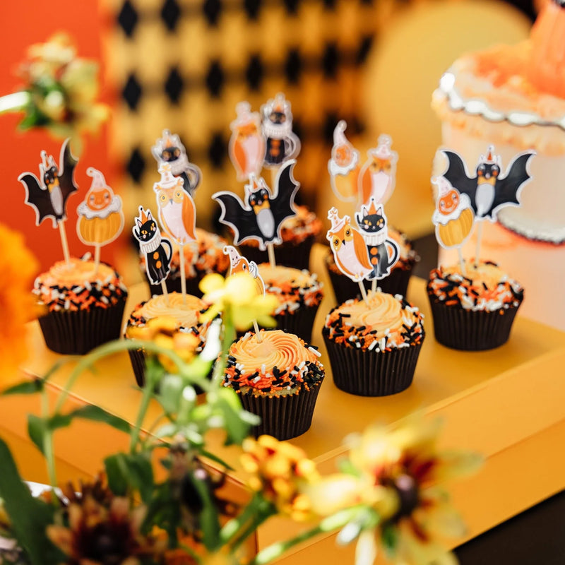 Tray of Halloween cupcakes with orange frosting and vintage character toppers on a yellow dessert table beside a cake