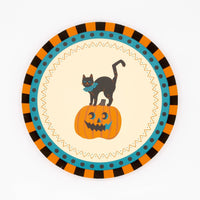 Vintage Halloween dessert plate with a black cat standing on a grinning jack-o'-lantern in a cream center, framed by orange-black striped and teal-dotted borders, overhead view on white background.