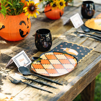 Black scalloped paper napkin with an orange owl in a party hat and foil stars under a pumpkin-shaped argyle plate at an outdoor Halloween place setting