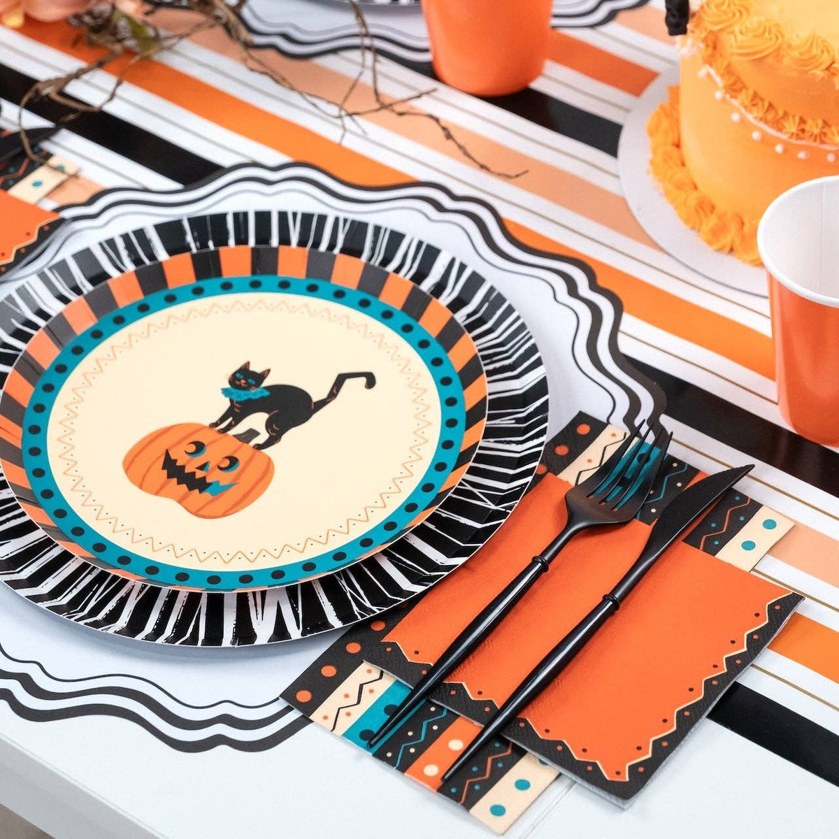 Styled vintage Halloween place setting showing the orange napkin with a black fringe rosette charger plate featuring a vintage black cat and jack-o'-lantern, gold flatware, on an orange-black-cream striped runner.