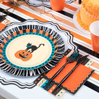 Styled vintage Halloween place setting showing the orange napkin with a black fringe rosette charger plate featuring a vintage black cat and jack-o'-lantern, gold flatware, on an orange-black-cream striped runner.