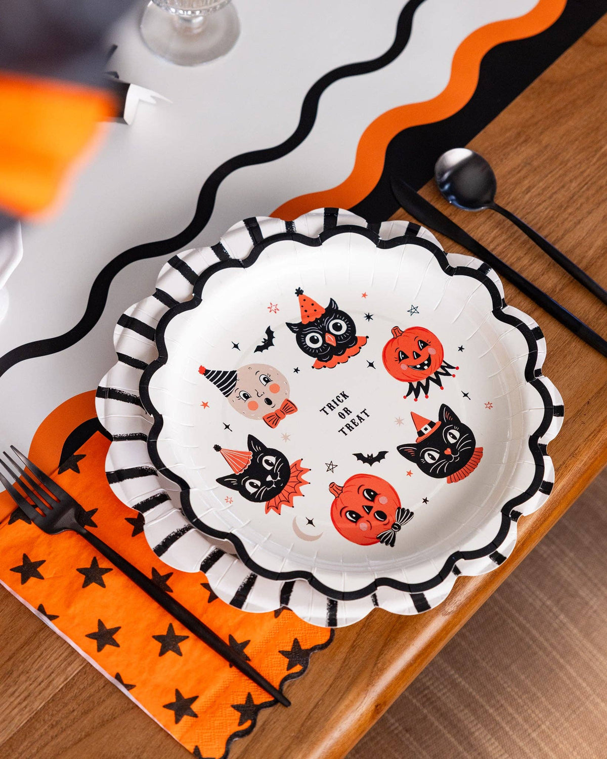 Halloween-themed plate with black cats, pumpkins, and moon on a wooden table paired with halloween themed napkins and table runner.
