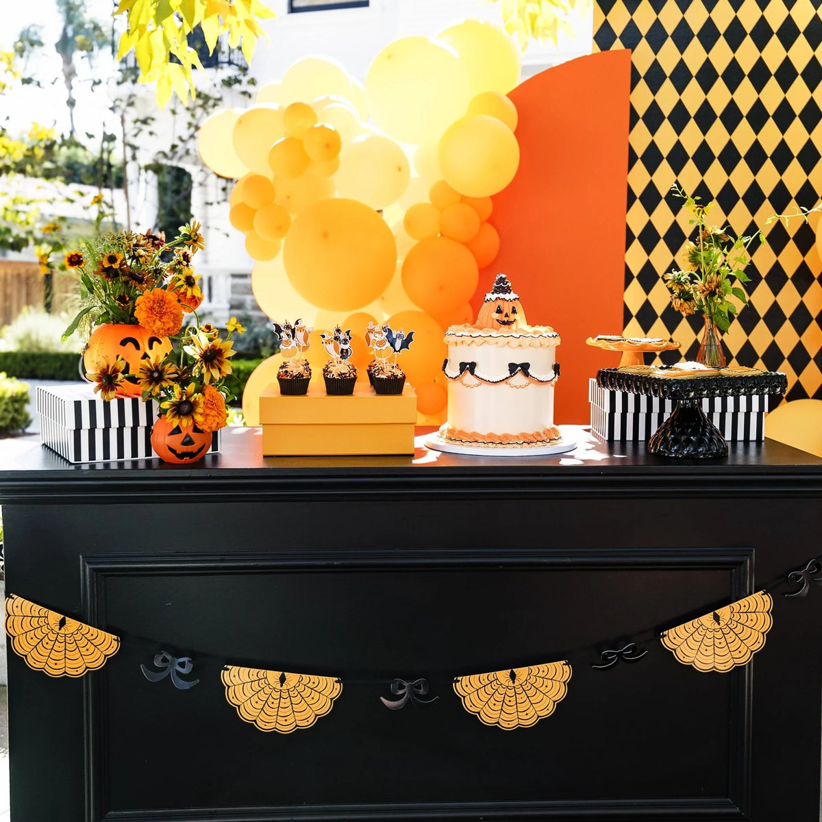 Orange spiderweb banner with black bows hung across a black dessert table with a jack-o'-lantern cake, cupcakes, and orange balloons
