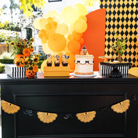 Orange spiderweb banner with black bows hung across a black dessert table with a jack-o'-lantern cake, cupcakes, and orange balloons