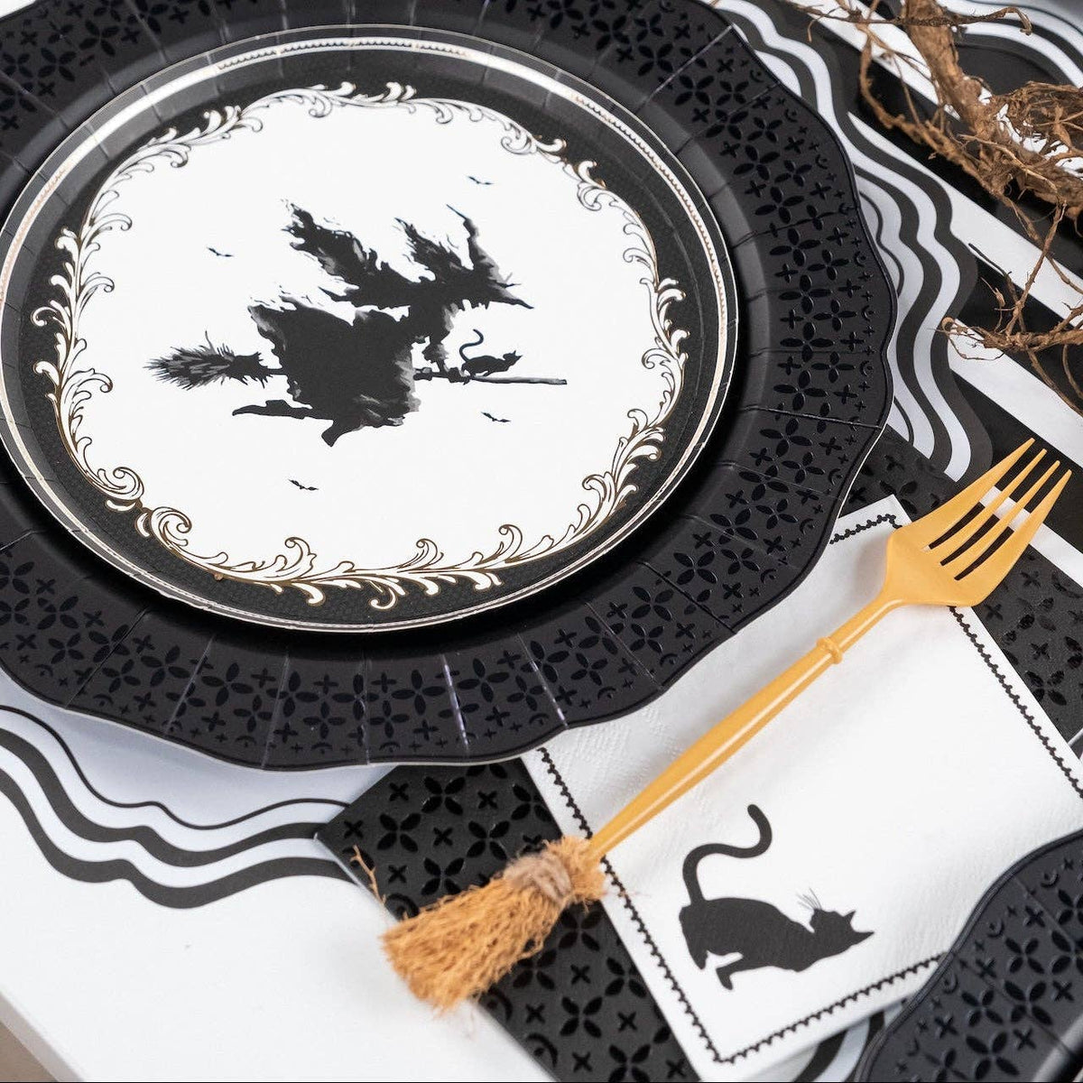 Layered Halloween place setting showing a black cat paper napkin tucked under a gold broomstick fork, resting on a black lace charger.
