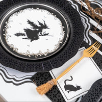 Layered Halloween place setting showing a black cat paper napkin tucked under a gold broomstick fork, resting on a black lace charger.