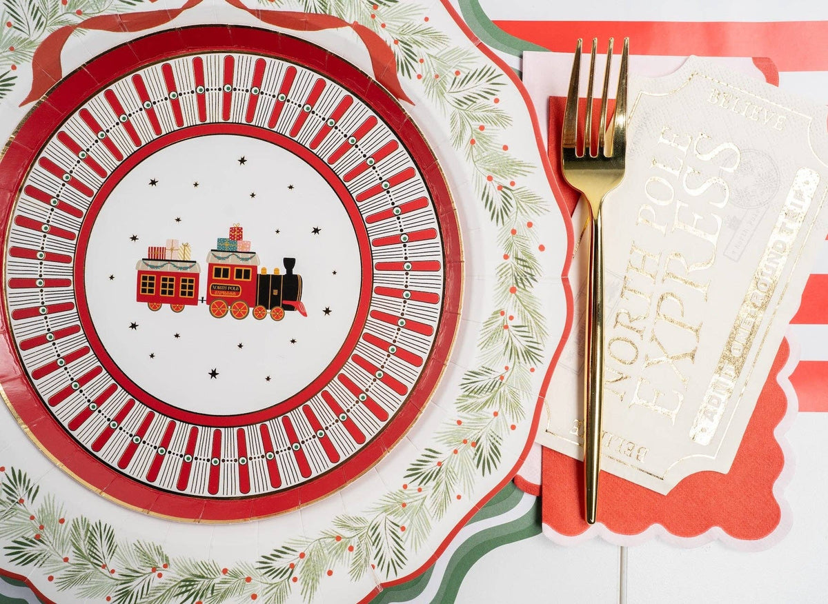 Place setting with a vintage red-train plate, holly rim, green scalloped charger, gold fork, and a cream guest towel with gold lettering.