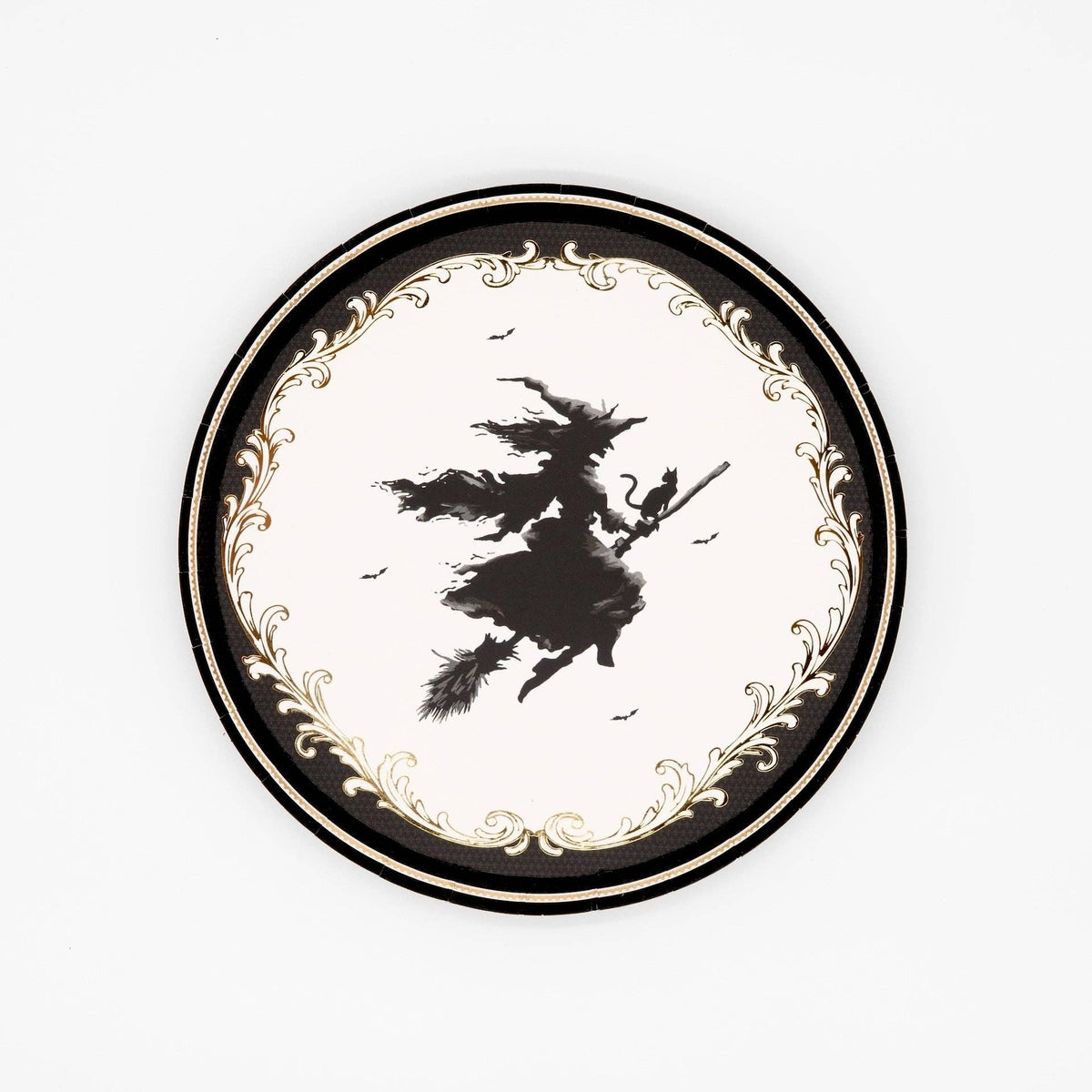 Top-down studio shot of a 7 inch round paper dessert plate featuring a vintage black flying witch silhouette and a metallic gold foil filigree border on a white background.