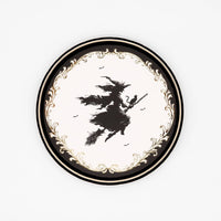 Top-down studio shot of a 7 inch round paper dessert plate featuring a vintage black flying witch silhouette and a metallic gold foil filigree border on a white background.