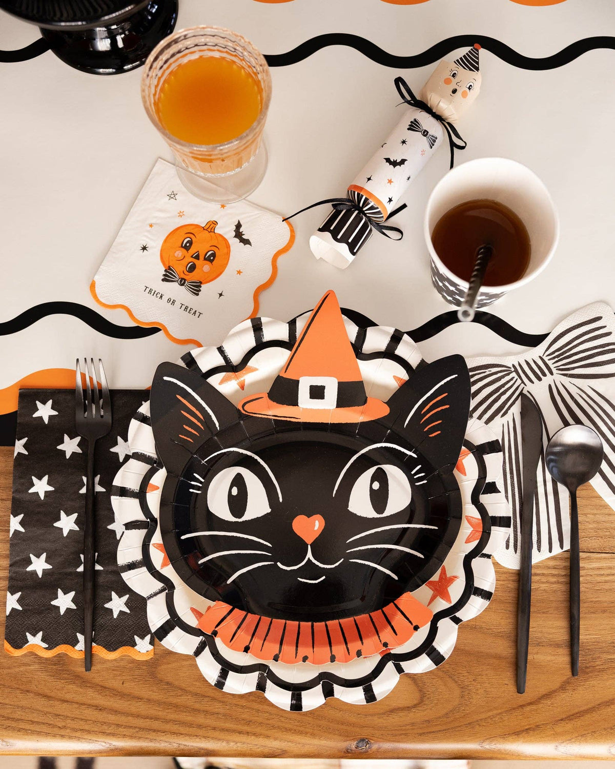 Halloween-themed table setting with witchy black cat plate, orange and black napkins, and drinks on a wooden table.