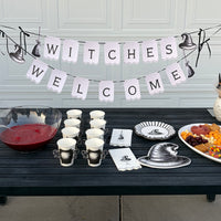 Full witch party table with the Welcome Witches banner, cauldron cups, black witch-hat plates, a red punch bowl, and orange snacks