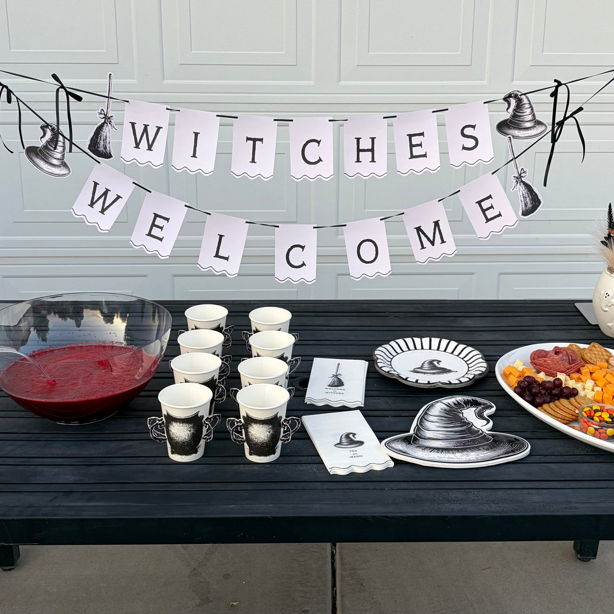 Full witch party table with the Welcome Witches banner, cauldron cups, black witch-hat plates, a red punch bowl, and orange snacks