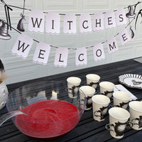 Witches Welcome banner over a black table with cauldron cups, black witch-hat plates, a red punch bowl, and black taper candles