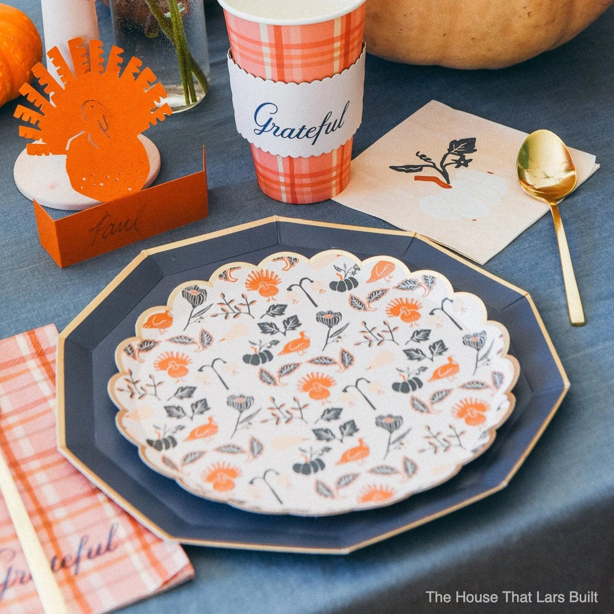 Close-up place setting with layered plates, plaid cup, and white pumpkin cocktail napkin next to gold flatware.