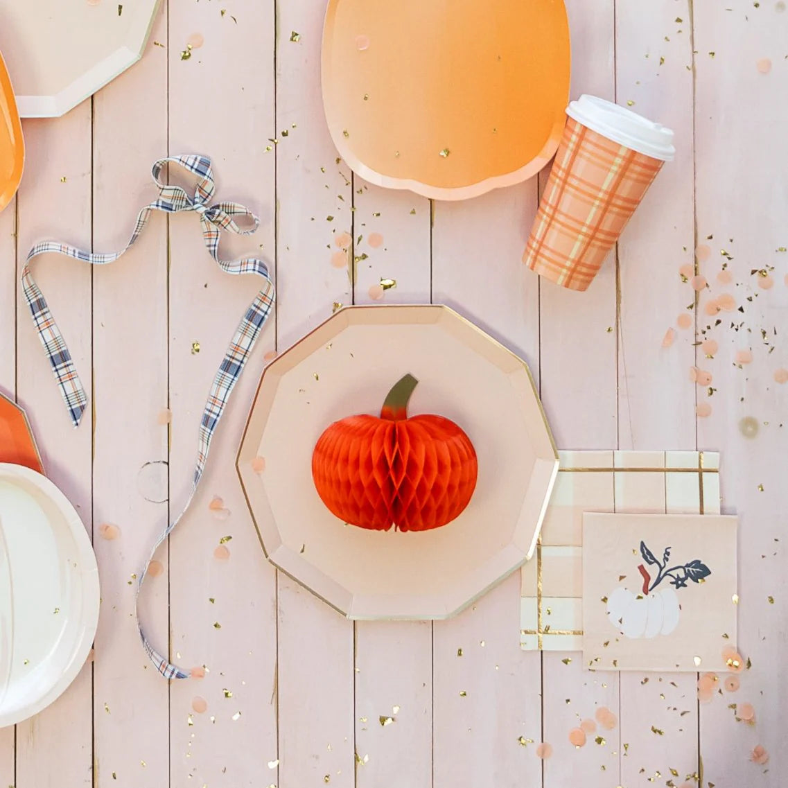 Flat lay of pastel pumpkin décor, octagon plates, plaid cup, and white pumpkin cocktail napkin with gold confetti.