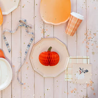 Flat lay of pastel pumpkin décor, octagon plates, plaid cup, and white pumpkin cocktail napkin with gold confetti.
