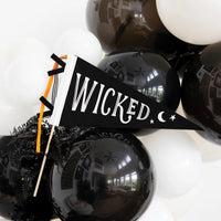 Hand holding black WICKED felt Halloween flag with moon and stars among balloons.