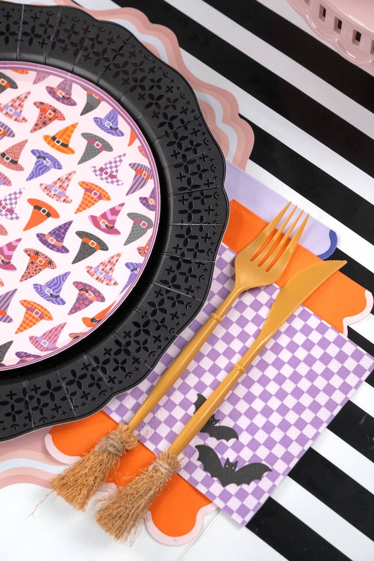 Detail shot of a single witch hat plate on a black charger — purple checkered napkin underneath, gold flatware alongside, showing the pattern and edge up close.