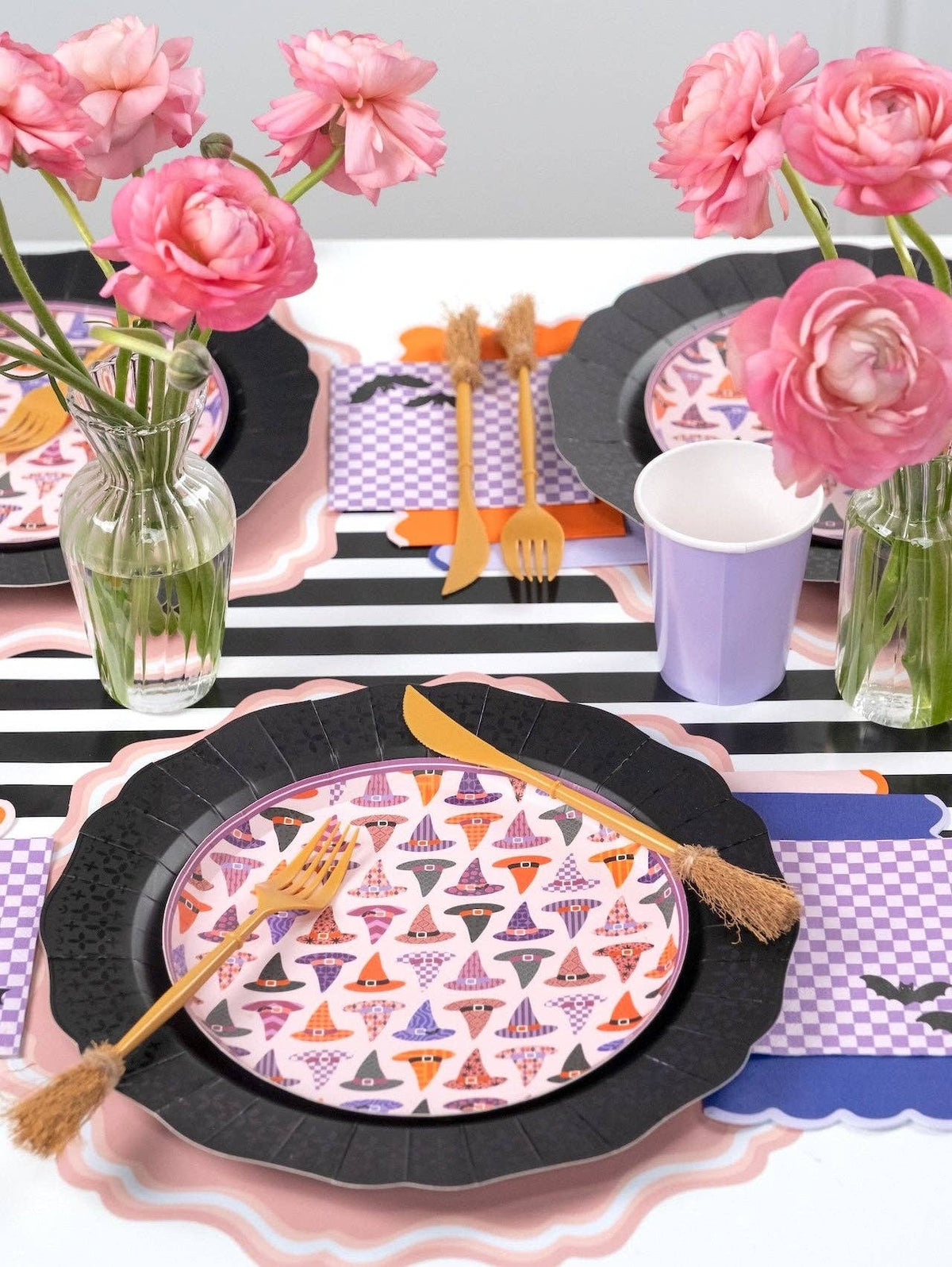 Top-down detail of two witch hat plate settings on black chargers with purple checkered napkins, gold flatware, and pink roses.